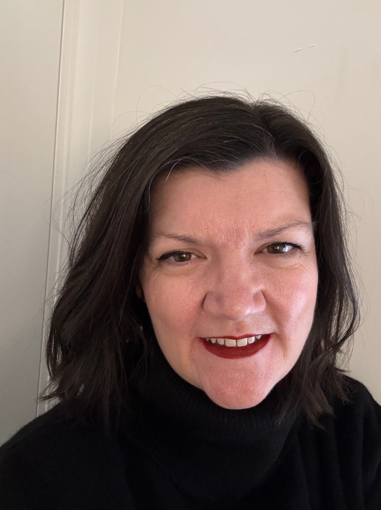 Close-up portrait of a woman with shoulder-length dark hair, wearing a black turtleneck. She has a friendly expression and is smiling slightly.